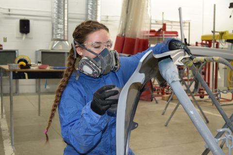 Female collision repair student wearing a respirator mask uses a sanding wheel to smooth a car fender's surface and prepare it for paint